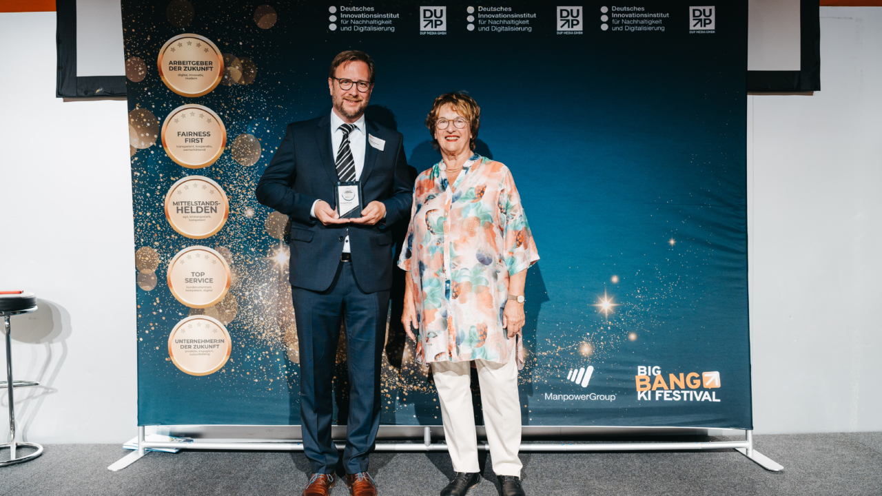 Zwei Personen bei der Preisverleihung, wovon eine den Award in der Hand hält. Vor einer blauen Wand mit Firmenlogos.